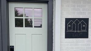 A light grey door with a keypad lock, a window with a view of trees, and a sign on the wall with house outlines and the text "EAST". - mi casa es TU casa (EAST) (Upland)