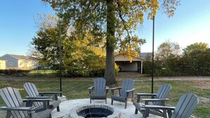 A fire pit surrounded by Adirondack chairs in a backyard with a lawn and a garage in the background. - mi casa es TU casa (Center) (Upland)