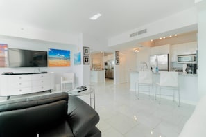 Uma sala de estar moderna com um sofá de couro preto, um móvel branco para entretenimento e uma mesa de centro de vidro. Há uma área de cozinha com armários brancos e um micro-ondas.