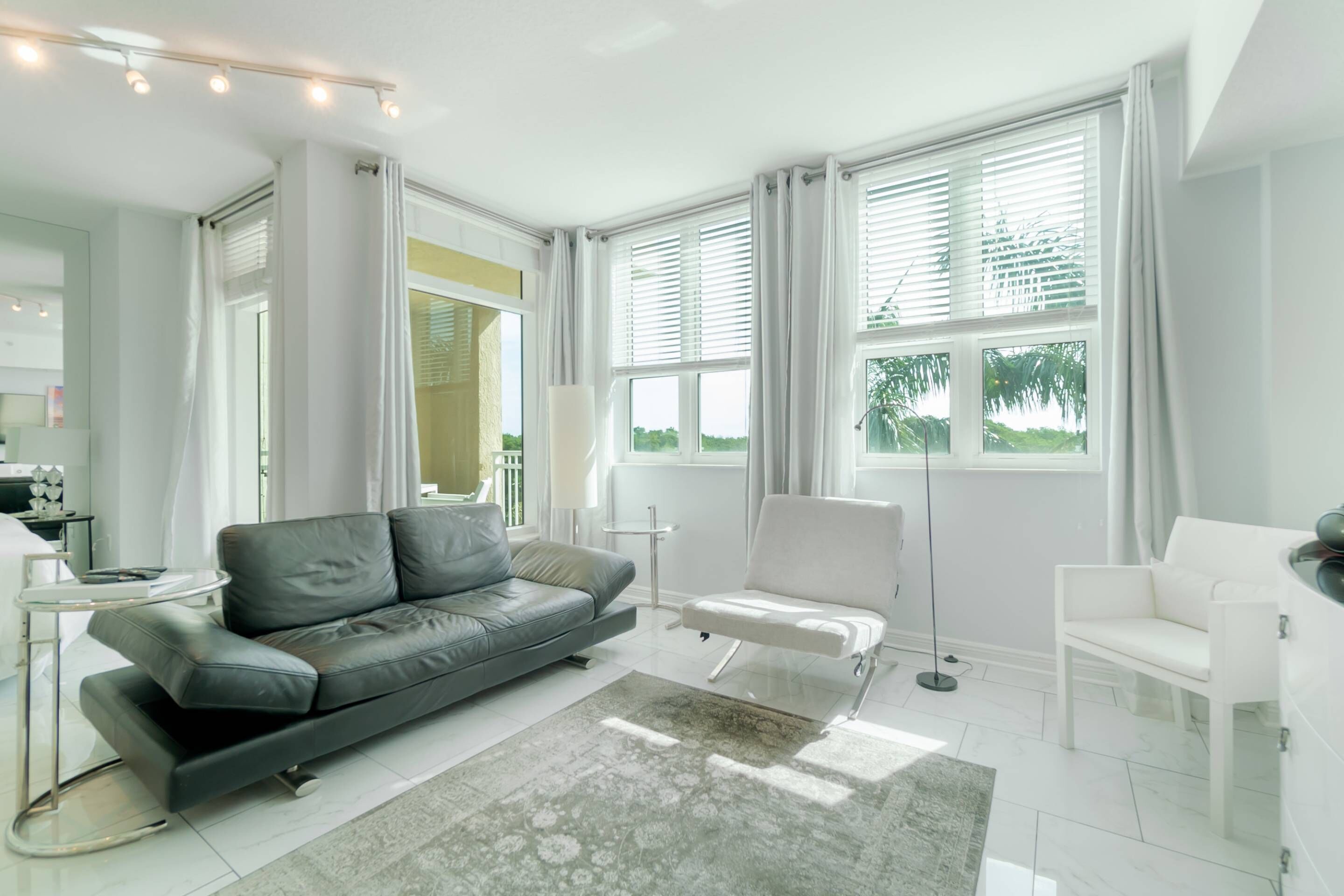 A modern living room with a grey sofa, a white armchair, and a coffee table. There are large windows with curtains, a lamp, and a view of greenery outside.