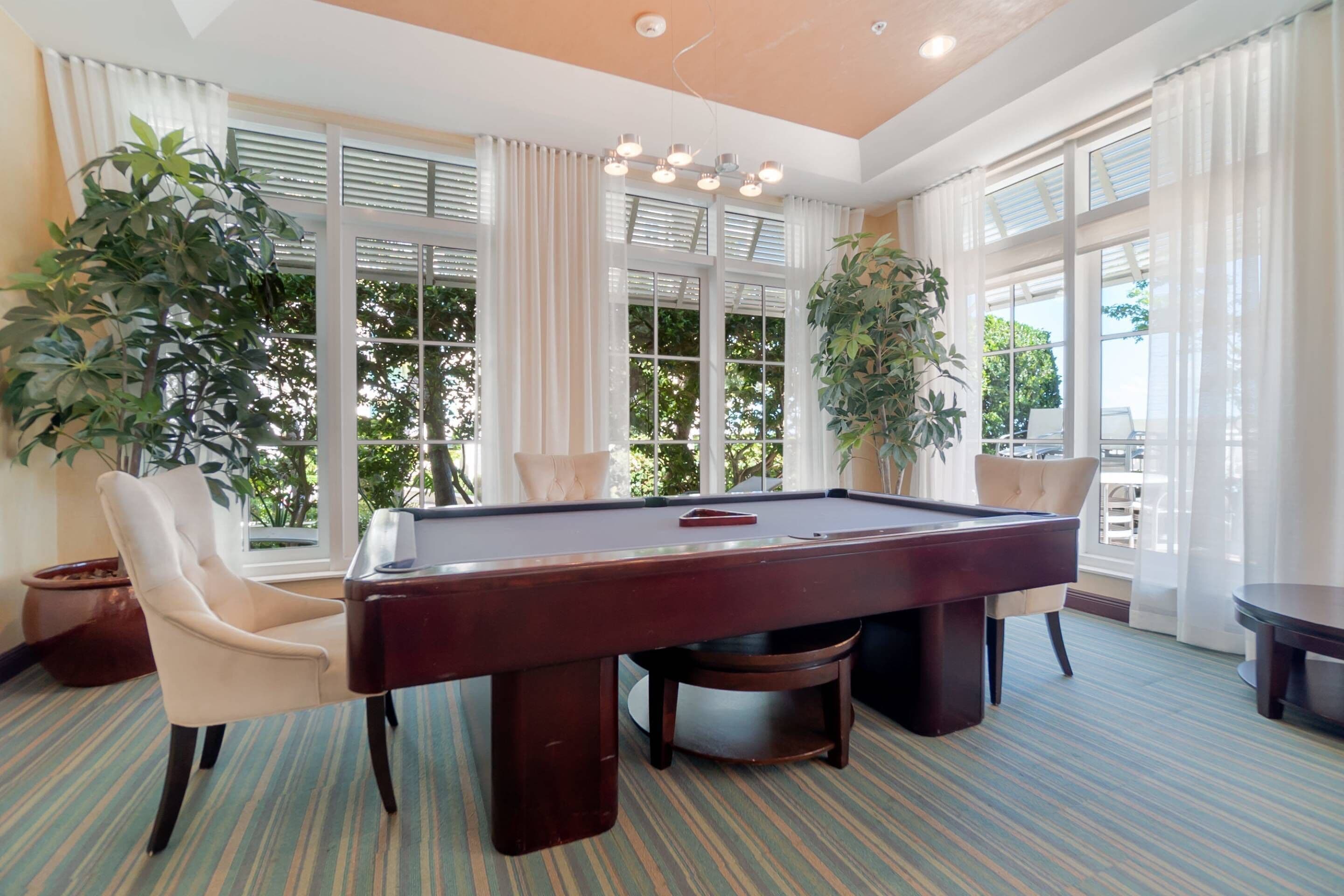 A pool table in a room with large windows, two chairs, and potted plants.