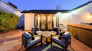 Un patio al aire libre con una mesa y cuatro sillas, una pequeña barra y una puerta de cristal que da a un espacio cubierto.