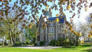 Ein großes, historisches Haus mit einem markanten Turm und zahlreichen Fenstern, umgeben von einem gepflegten Rasen und Bäumen.