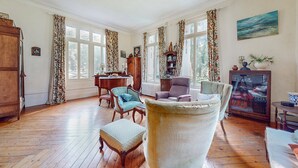 Ein geräumiges Wohnzimmer mit Holzböden, einem Flügel, einem bequemen Sessel und einem großen Fenster mit floralem Vorhang.