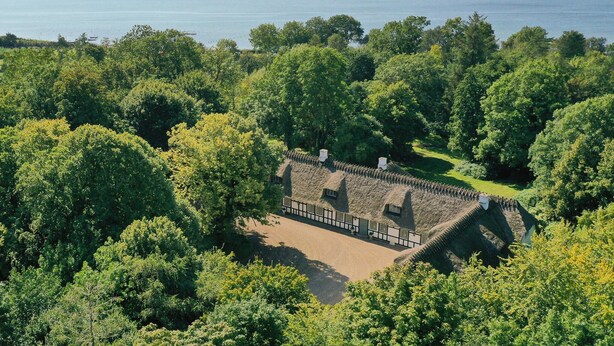 Un bâtiment à toit de chaume, entouré d’arbres denses, avec une vue sur une vaste étendue d’eau en arrière-plan.