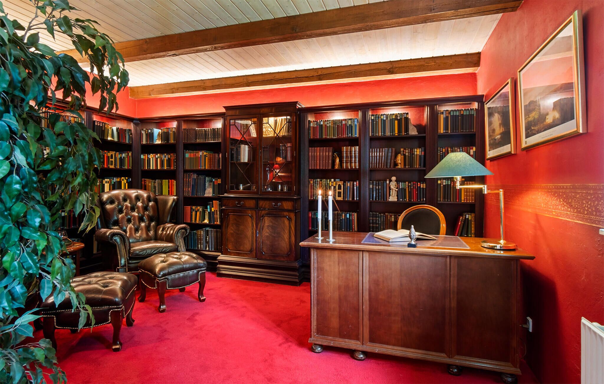 Une bibliothèque avec des fauteuils en cuir, des étagères, un bureau avec une lampe et un tapis rouge.