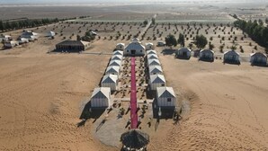 Vogelperspektive eines Wüstenresorts mit mehreren Jurten, die entlang eines zentralen, roten Weges angeordnet sind, umgeben von spärlicher Vegetation und Gebäuden in der Ferne.