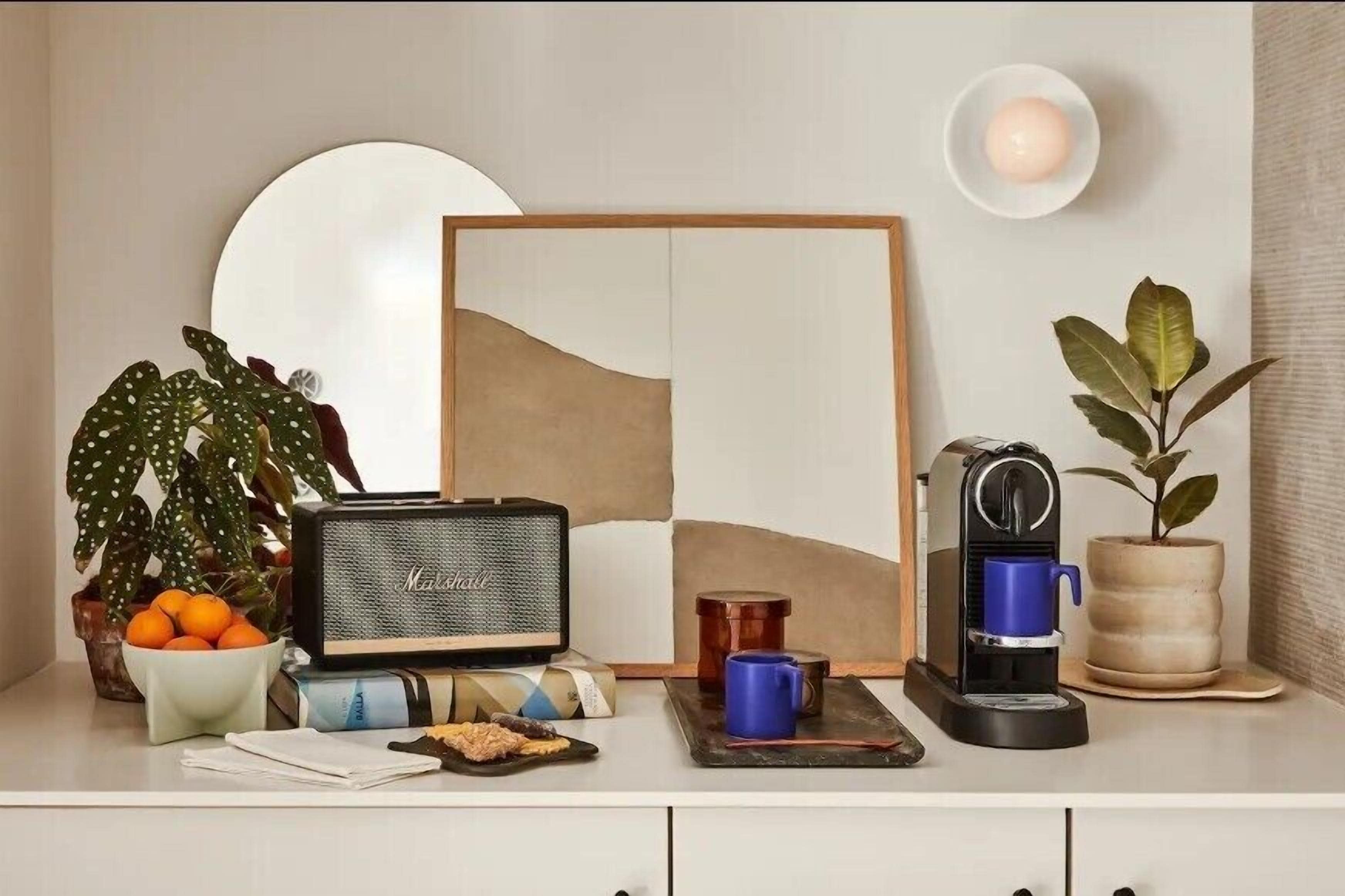 A neatly arranged coffee station with a coffee maker, a bowl of oranges, a potted plant, and a framed artwork on a white counter.