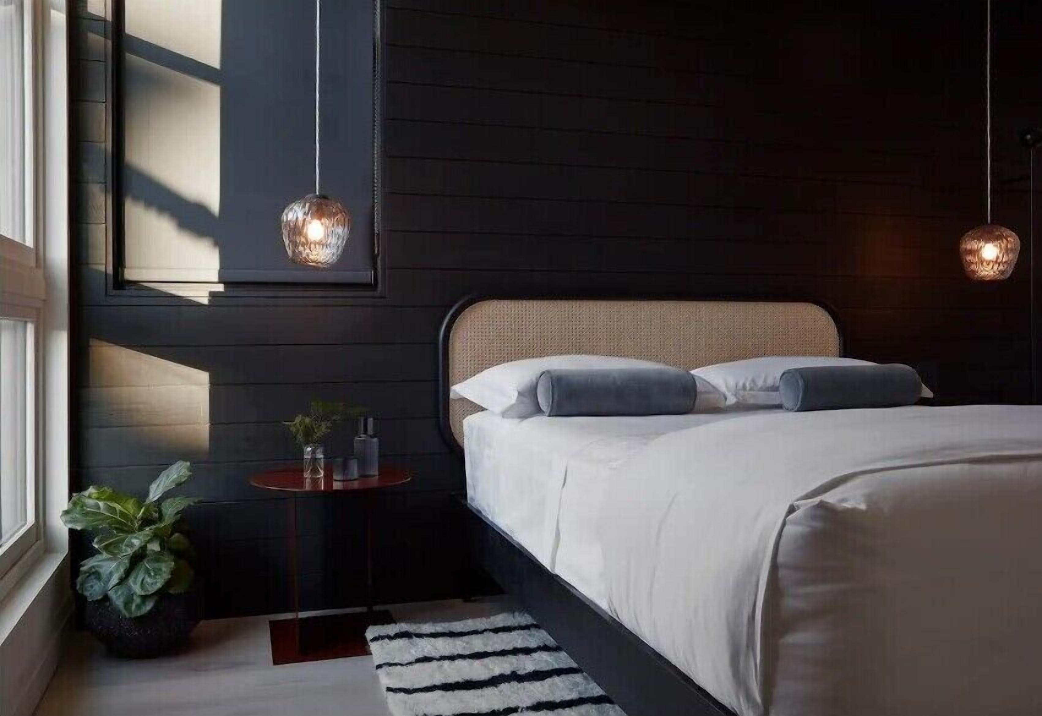 A modern bedroom with a large bed, a small bedside table, a potted plant, and two hanging pendant lights.