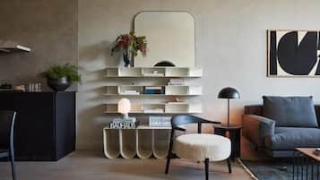 A modern living room with a minimalist design, featuring a black sofa, a round ottoman, a black armchair, a side table with a lamp, and a bookshelf with decorative items.