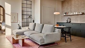 A modern living room with a grey sectional sofa, a wooden coffee table, and a pink rug. There is a dining area with a table and chairs, and a kitchen area with a sink and shelves.