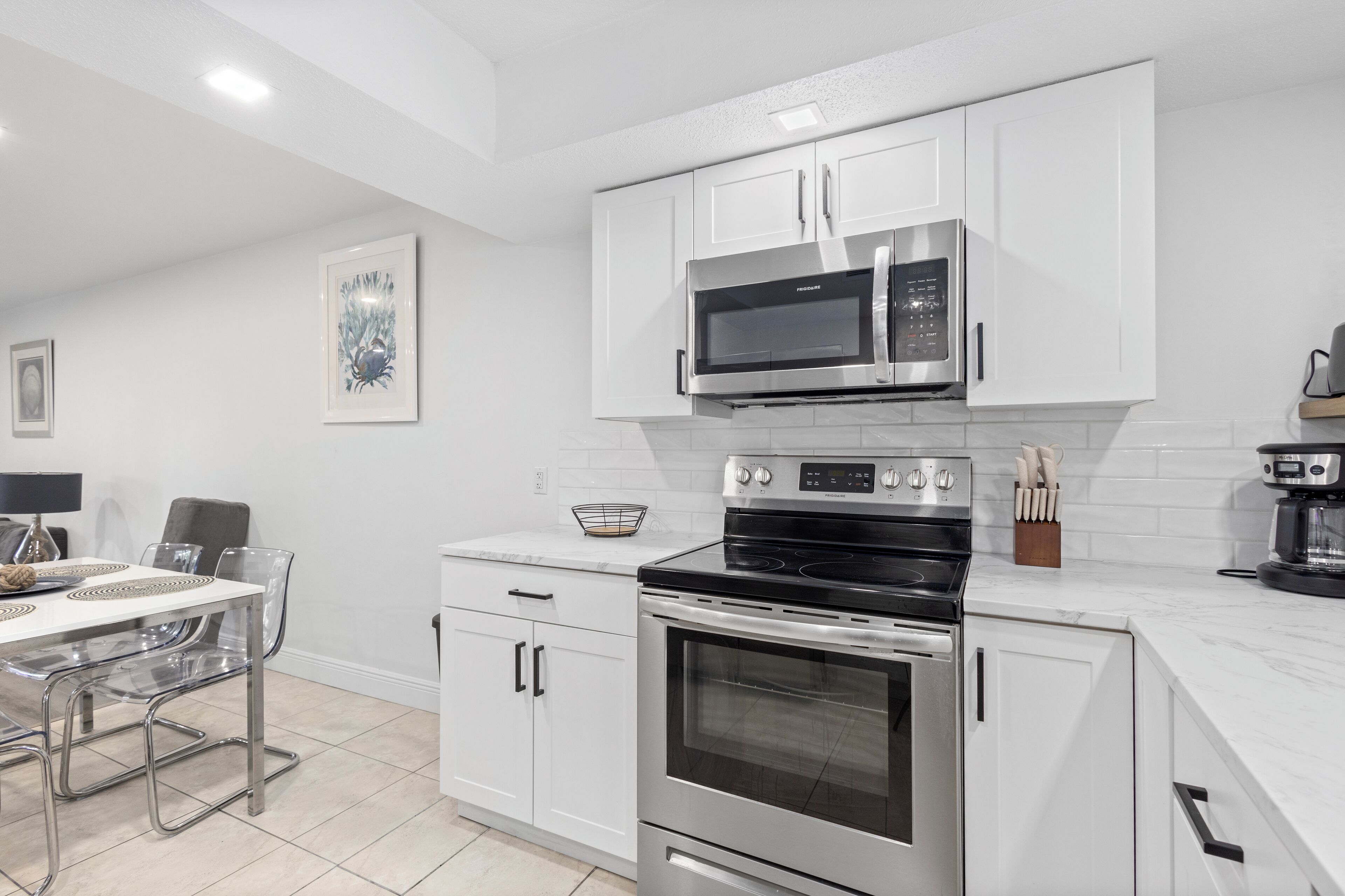 A modern kitchen with white cabinetry, stainless steel appliances, and a dining area with clear chairs.