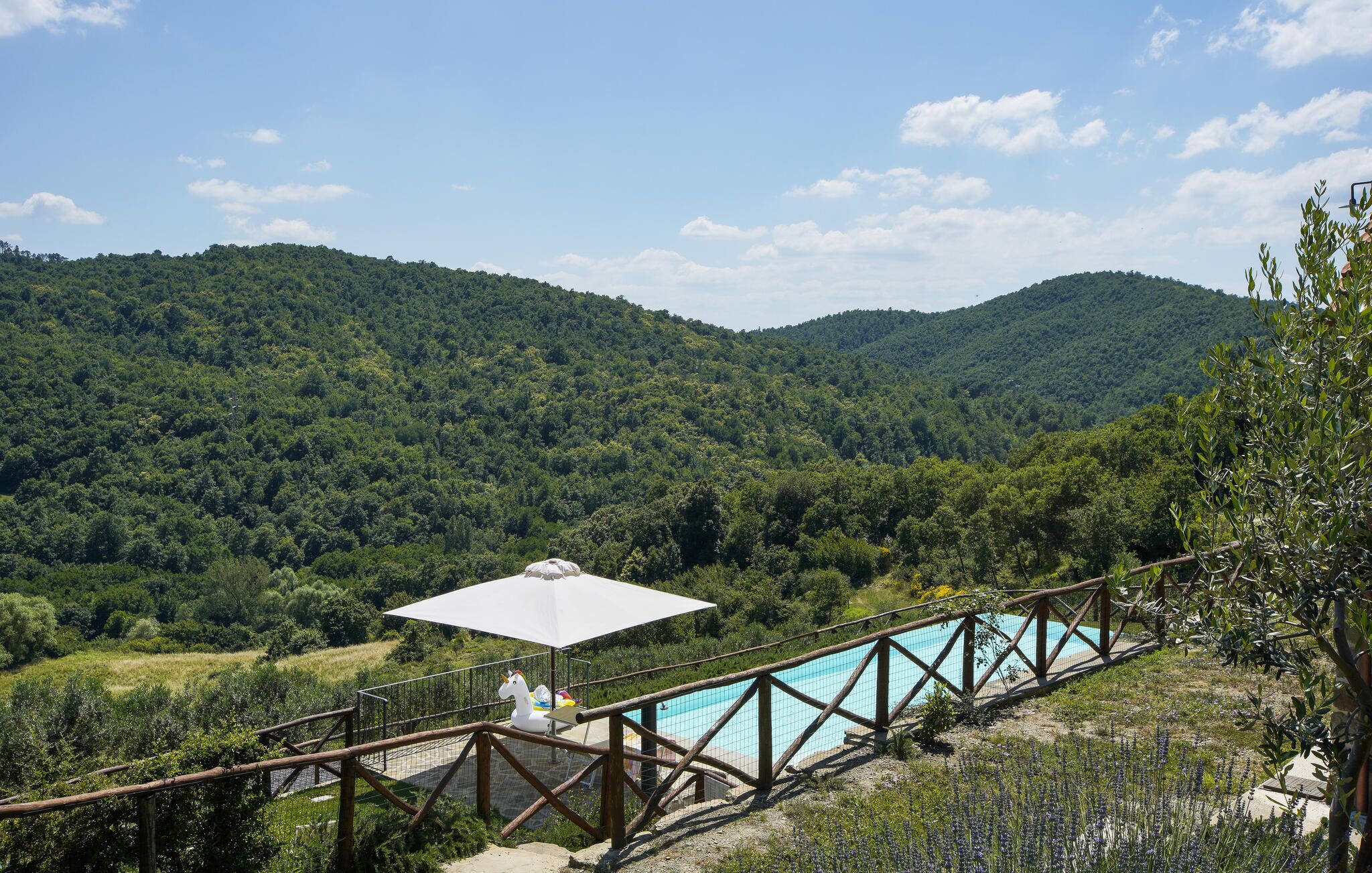 A pool area with a wooden deck and a white umbrella, surrounded by lush green hills.