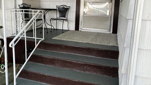 Una escalera lleva a una puerta con el número 136 arriba, una pequeña mesa con sillas, y una ventana con vistas a árboles.