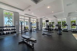 Uma academia ampla, com diversos equipamentos de exercício, janelas grandes e um banco.