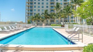 A rectangular swimming pool with clear blue water, surrounded by white lounge chairs and palm trees, with a multi-story building in the background.
