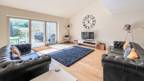A spacious living room with a large TV, comfortable seating, a clock, and a view through the glass doors.