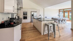 A modern kitchen with white cabinetry, stainless steel appliances, and a wooden floor. There is a dining area with a table and chairs, and a large window providing natural light.