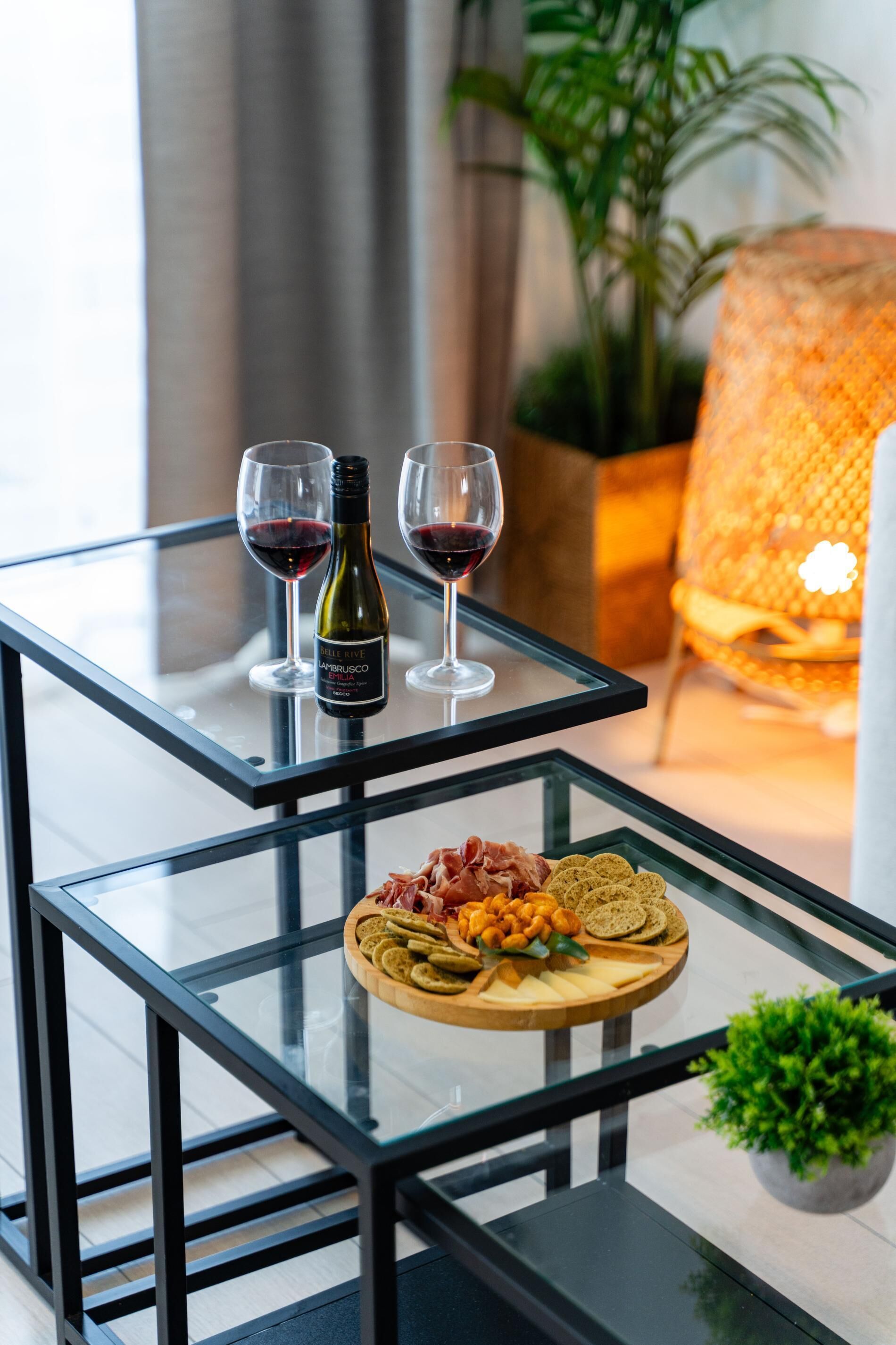 Ein Tisch mit Glasplatte, zwei Weingläsern und einer Weinflasche, ein Holzbrett mit Käse und Crackern sowie ein Blumentopf.