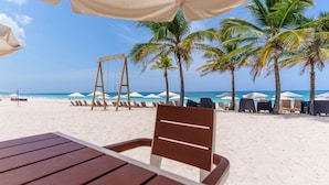 A beach with a wooden table and chairs under a canopy, palm trees, and beach umbrellas.
