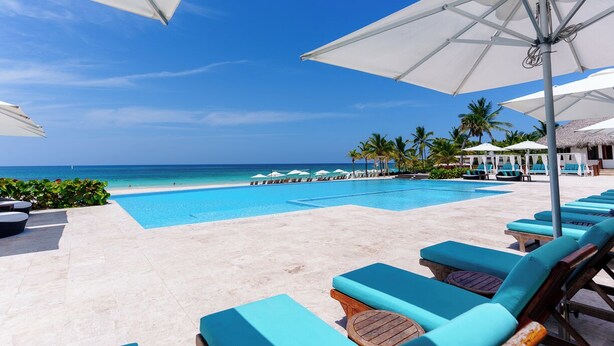 Un espace aménagé au bord de la piscine, avec des chaises longues et des parasols, une vue dégagée sur l’océan et une plage de sable.