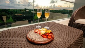 Un balcón con una mesa dispuesta para dos personas, que incluye un plato de comida y dos copas de vino, con vistas a un paisaje pintoresco.