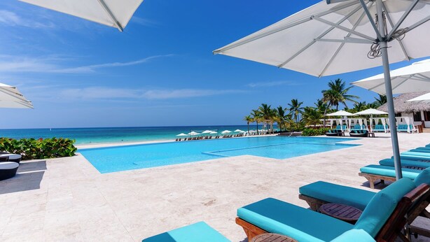 Un espace aménagé au bord de la piscine, avec des chaises longues et des parasols, offrant une vue dégagée sur l’océan, le tout dans un cadre tropical agrémenté de palmiers.