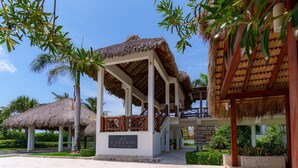 Un complexe hôtelier avec des bâtiments à toits de chaume, une enseigne indiquant le «Canaba Bay Beach Club», et un ciel d’un bleu limpide.