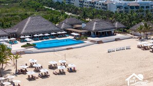 A resort with a large swimming pool, beachside lounge chairs, and thatched-roof structures.
