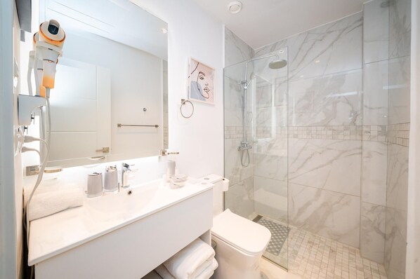 Un baño moderno con un tocador blanco, un espejo grande, un inodoro y una zona de ducha con mampara de vidrio.
