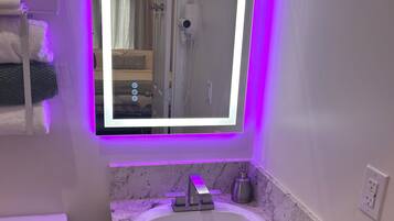 Un bagno con lavandino bianco, uno specchio con illuminazione a LED viola, un water e un portasciugamani.