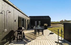 Una terraza de madera con una mesa, sillas, una parrilla y un cobertizo pequeño al fondo.