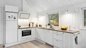 A modern kitchen with white cabinetry, a built-in oven, a sink, and a window overlooking greenery. - Beautiful home in Blokhus with sauna (Blokhus)