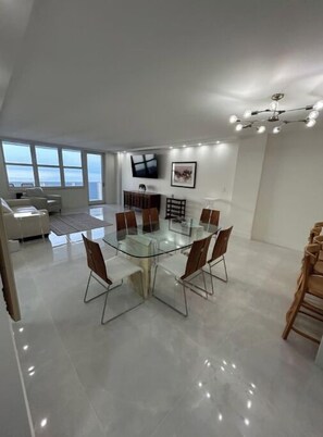 A modern dining area with a glass table surrounded by chairs, a flat-screen TV mounted on the wall, and a ceiling light fixture. - Panoramic Beachfront Views! (Hollywood)