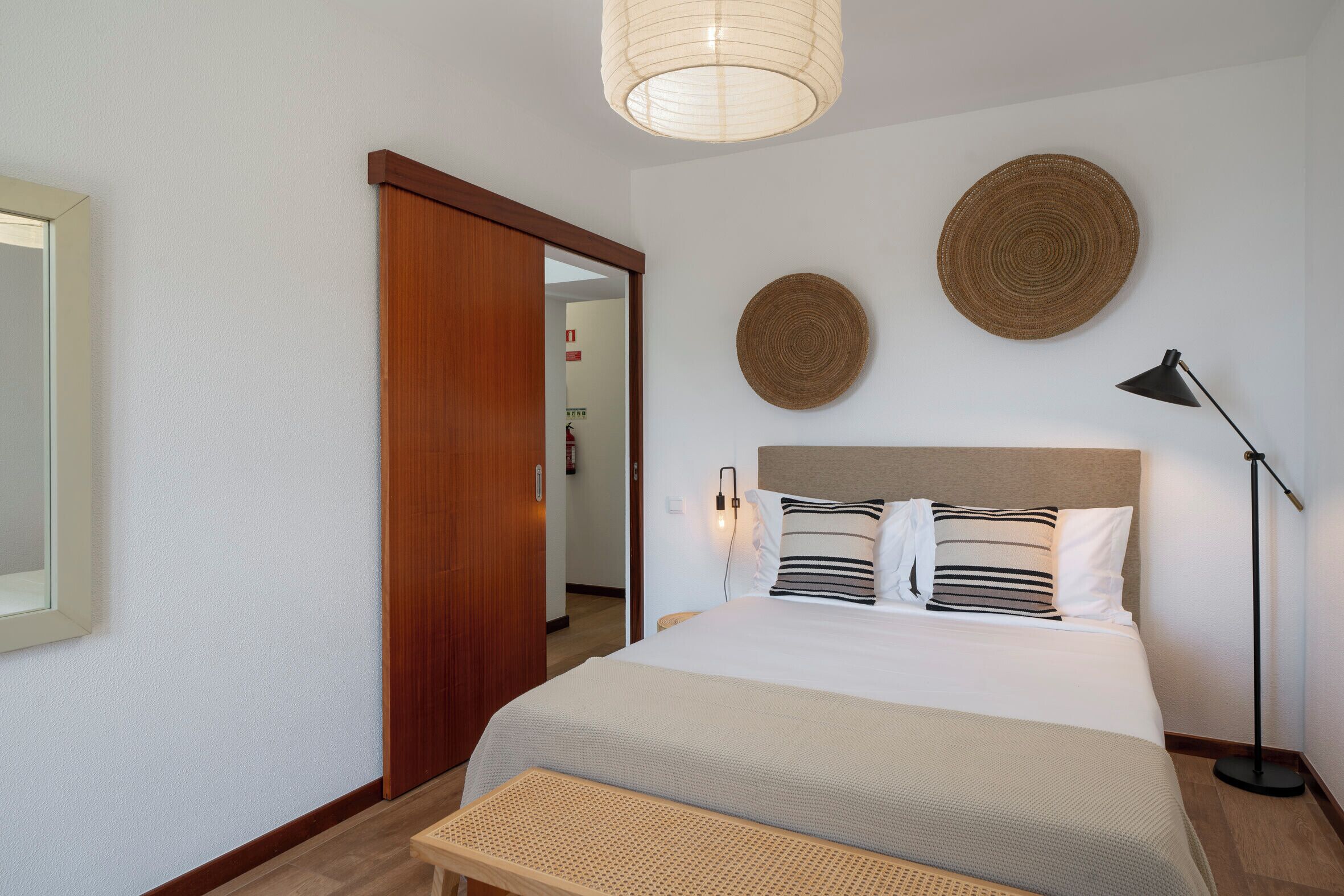 A hotel room with a bed, a bedside lamp, a woven bench, a large mirror, and two woven wall decorations.