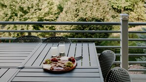 Une table dressée avec une planche de charcuterie, deux verres contenant une boisson claire et une vue sur un jardin avec des arbres et une balustrade.