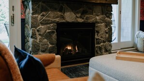 Une cheminée en pierre avec un feu de bois, un canapé avec des coussins et une fenêtre donnant sur un beau paysage.