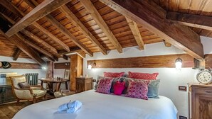 Ein Schlafzimmer mit Holzbalkendecke, einem Bett mit vielen Kissen, einem Nachttisch und einem Sessel.