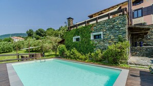 Ein rechteckiger Pool mit umlaufender Holzterrasse, vor einer Steinsäule und üppigem Grünwerk.