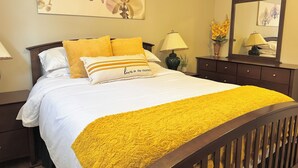 Una cama bien hecha con una manta amarilla y almohadas, una lámpara de noche, un espejo y una obra de arte floral en la pared.