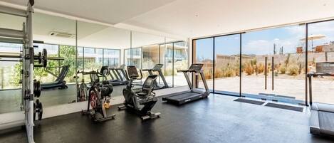 A modern gym with cardio machines, a weight bench, and large windows offering a view of the outdoors.