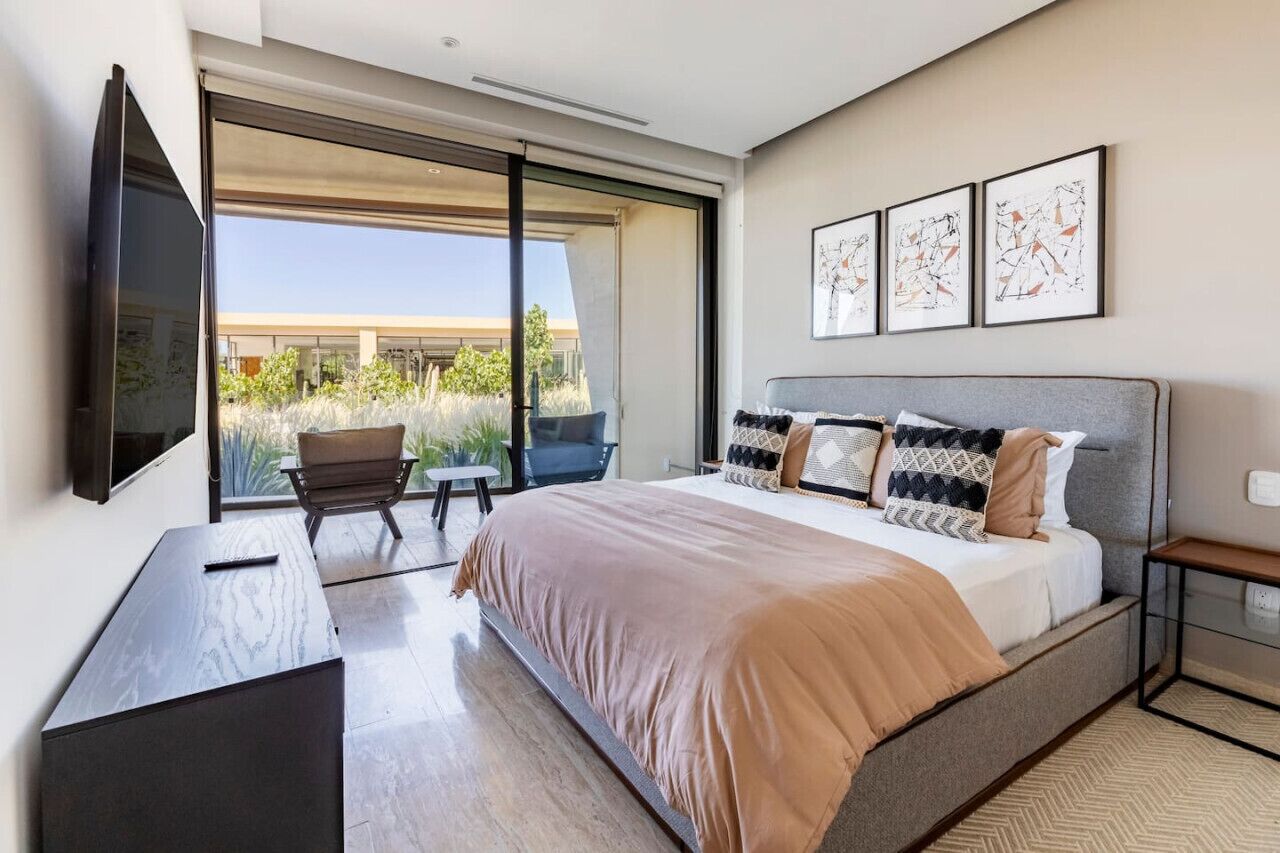 A modern bedroom with a large bed, a flat-screen TV, a desk, and a chair. There are three framed artworks on the wall and a view of greenery outside.