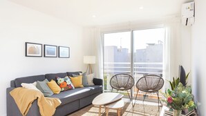 Un soggiorno moderno con un divano grigio, un tavolino in legno, due sedie nere, una grande finestra con vista sugli edifici e un vaso con fiori su un comodino in legno.