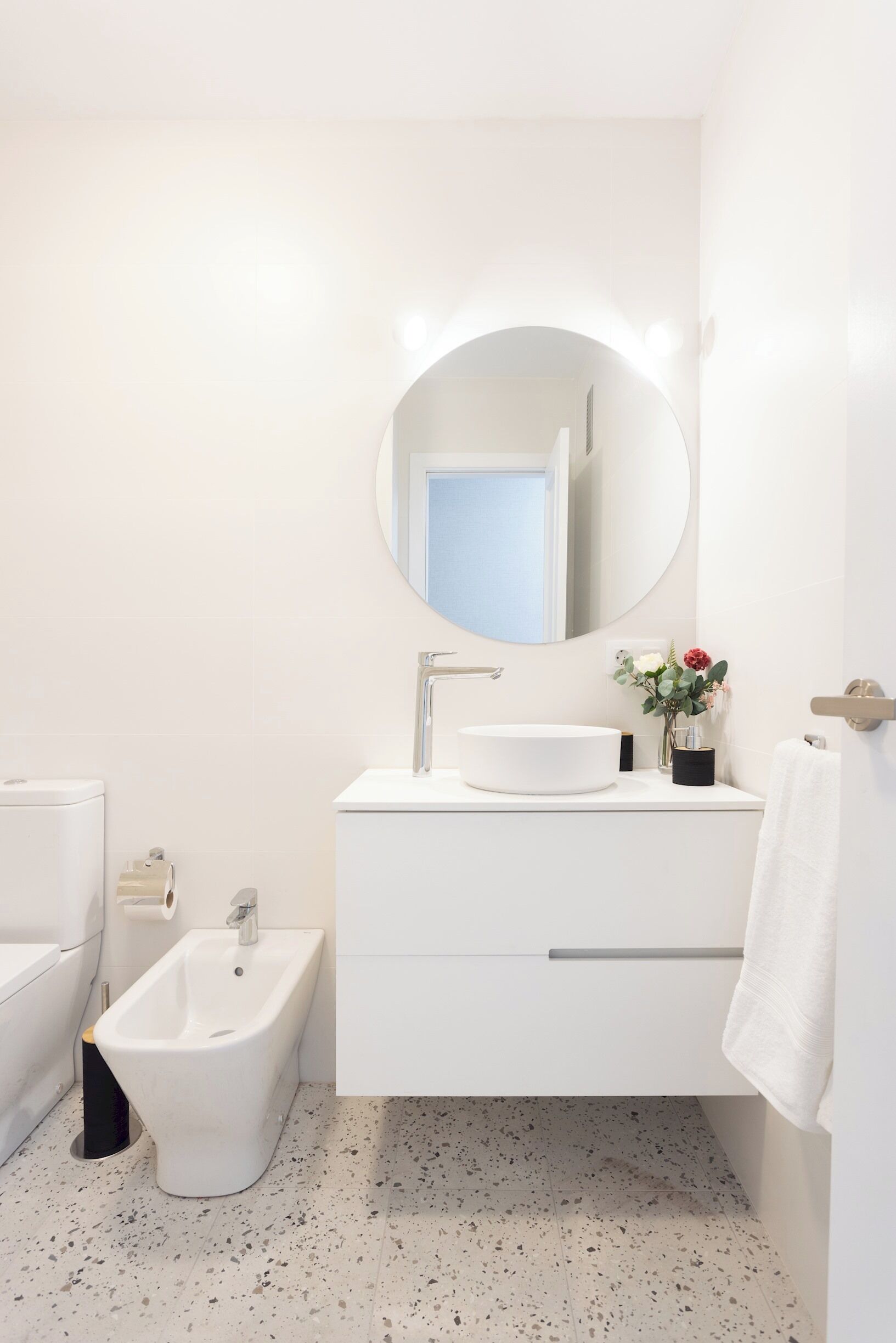 Ein modernes Badezimmer mit rundem Spiegel, weißem Waschbecken, Bidet und Handtuchhalter.