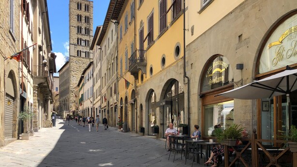 Eine schmale Straße, gesäumt von historischen Gebäuden, einige mit Balkonen und Bögen, und mit Außenplätzen eines Cafés.