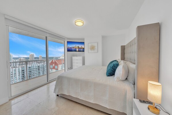 A bedroom with a large bed, a nightstand, a lamp, a painting, and a view of a cityscape through large windows.