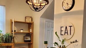 Una zona de menjador amb una taula de fusta, cadires verdes, una làmpada penjant, un rellotge i un cartell a la paret que diu "EAT".