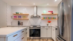 Una cocina moderna con armarios blancos, un horno de acero inoxidable y un refrigerador. Hay estantes de madera con utensilios de cocina y un letrero de neón con forma de cactus.