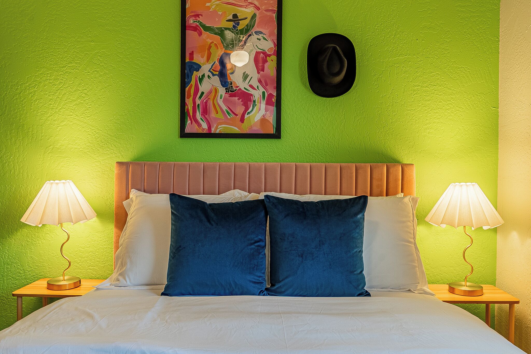 Une chambre d’hôtel avec un mur vert, un tableau encadré, un chapeau noir, deux lampes de chevet, un lit avec des oreillers blancs et bleus, et une tête de lit en bois.