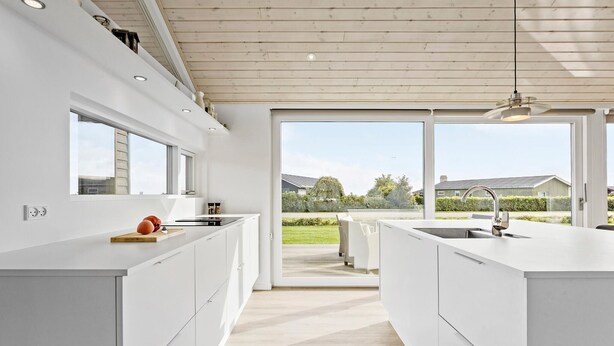 A modern kitchen with white cabinetry, a central island, and large windows offering a view of the outdoors. - Cozy home in Sæby with house sea view (Sæby)